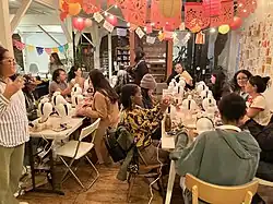 Two long tables of women-identifying and non-binary community members underneath hanging lanterns and papel picado.