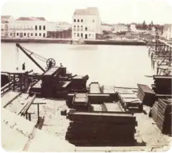 An old photograph showing piles of construction materials and equipment along the bank of a river with large white buildings lining the opposite bank