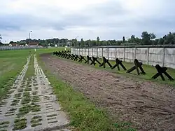 Strip of bare ploughed earth flanked by a concrete road on one side and a row of barricades and a fence on the other side, with buildings visible in the far background