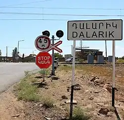 Image of a sign bearing the name of the village in English and Armenian. Just beyond the sign is a road that crosses a train track, with a stop sign in front of the tracks warning drivers of the passing train.