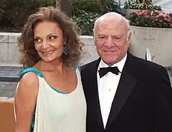 A woman with wavy and curly hair wearing a white dress lined with a teal fabric smiles as she looks to her right while standing next to a man wearing a tuxedo