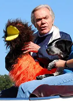 Dick Goddard is sitting in a car, holding a large brown-and-orange woolly bear caterpiller puppet in his hands. Next to him is a small dog being held by an assistant off-camera.