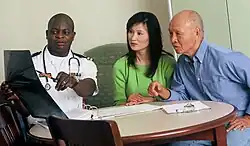 A black male doctor wearing a Public Health Service uniform is pointing at an X-ray image as he explains it to an elderly East Asian male and an adult East Asian female.