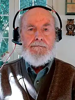 A grey-bearded man at home wearing a wired headset staring directly into the camera, as if on a remote meeting.