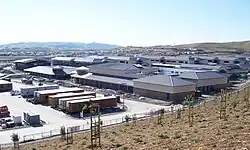 Several school buildings, nearly finished but with exposed cement walls where stucco will be. The sidewalk and road in front are also incomplete, and gravel is found in their place. The parking lot in front is full of trailers and trucks used by construction personnel.
