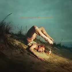 A photo of Eartheater lying on a beach on her stomach with her legs backwards over her head, with a small ornament dangling from her toes