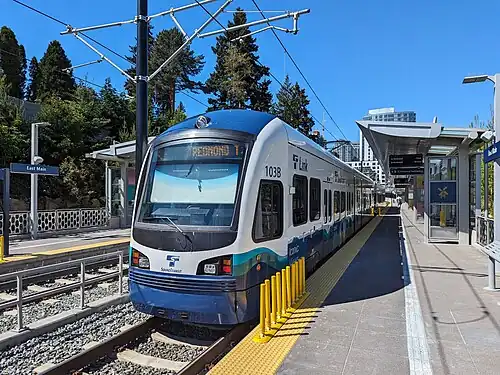 A modern light rail train, mostly white with blue accents, is stopped at the eastbound platform at East Main station in May 2024. The station has two platforms on the sides of the station with two tracks running down the center. Behind the train, there's a station with a covered platform and some greenery. The sky is clear and blue.