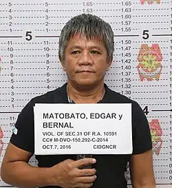 Chest-length shot of Matobato standing in front of a height chart, holding a paper with his name