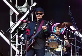 Edmund Shklyarskiy of Picnic wearing a dark shirt, pants, and cap, with black sunglasses, playing electric guitar on an outdoor stage, with drummer behind him