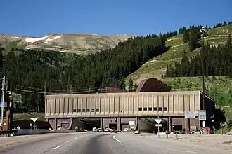 Automobiles are driving on a road leading to one of two openings in a building against a mountain. Letters above each opening read "Johnson Tunnel 1979" and "Eisenhower Tunnel 1973". On the roof of the building, large ventilation hoods are visible.