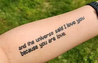A pale white arm, tattooed with the quote "and the universe said I love you because you are love", all lowercase and in a Minecraft-like font.