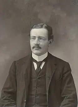 A mustachioed man with slick hair and round glasses in a black suit with a tie and popped collar
