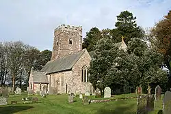 Stone building with square tower. Trees are to the right and behind with gravestones in the foreground.