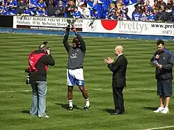 Four men standing on a grass football pitch. The man second from left, wearing a grey top, white shorts and white socks, is holding a trophy above his head. Spectators wearing blue or black tops are visible in the background.