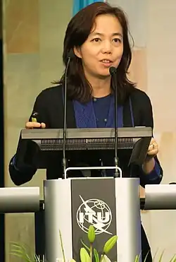 Li speaking at a conference, standing at a podium with the AI for Good logo visible on the podium