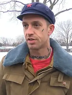 A photograph of a man wearing a flat cap with a red star on the front and a fur-lined jacket with a red star pinned to the left chest. "KPACABA" is tattoo'd to the man's neck.