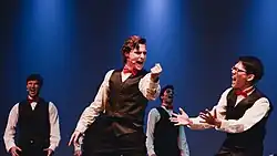 Four young men in tuxedoes sing on stage in front of a deep blue background. Two singers in particular stand in the foreground, gesticulating while singing a big note.