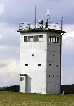 A squat, square white tower on a sloping meadow. Each side of the tower has an unbroken row of windows near the top. There is a searchlight on the tower's flat roof, poles carrying equipment and a railing around the roof's outer edge.