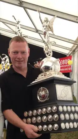 Short haired man carrying with both arms a large silver coloured trophy having a polished wood plinth