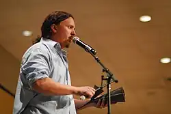 Donovan onstage, speaking into a microphone and hitting a cowbell