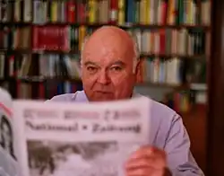 An image of Gerhard Frey reading the newspapers with a backdrop of books