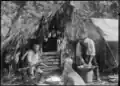 Two men washing dishes in a basin in front of a shelter thatched with palm fronds.