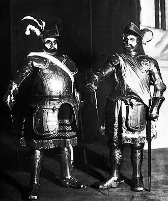 Two men standing in full armor. They have on hats with feathers and are carrying swords