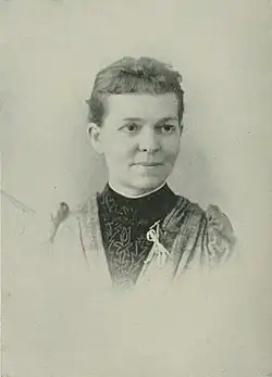 B&W portrait photo of a woman with her hair in an updo, wearing a clergy collage, dark bloues, and light jacket with a white ribbon pinned upon it.