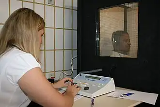 a female medical professional is seated in front of a special sound-proof booth with a glass window, controlling diagnostic test equipment. Inside the booth a middle aged man can be seen wearing headphones and is looking straight ahead of himself, not at the tester, and appears to be concentrating on hearing something