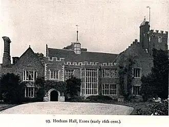 Black and white photograph of H-plan red brick tudor house with a porch on the left, two decorative oriels on the right, and a tall Tudor tower in the background connected to the building.