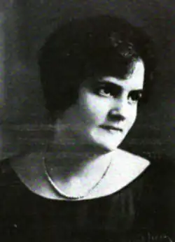 A woman with light skin and dark hair and eyes, wearing a dark top and a strand of pearls