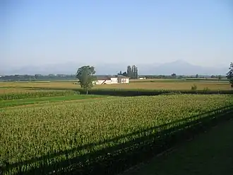 Photo of a rural scene with a farmhouse and crops in the foreground and mountains in the distance.