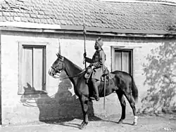 In a black and white photograph, a man wearing a turban and military uniform sits astride a stationary, dark-coloured horse, facing left. In his right hand he holds aloft a sword. Directly behind them stands a single-storey, brick building with two large windows and a tiled roof.