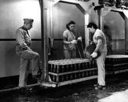 Photograph of a man in a military uniform observing two young women working at a rack of mortar shells