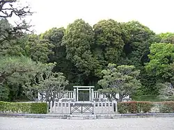 Concrete torii gate behind two concrete fences and in front of a group of trees.
