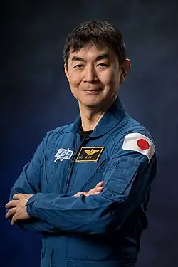 JAXA astronaut Kimiya Yui, an Asian man in a blue flight suit, is in front of a dark background.