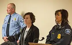 A police officer in uniform, a mayor in a black blazer, and a police chief in a dark blue police jacket stand before a podium.