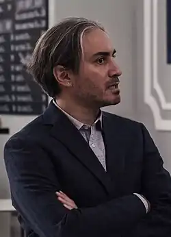 white caucasian male in suit, around 30 years old, appears concerned with arms crossed, dark hair with fine blond highlighted fringe