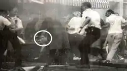 low-resolution monochrome image showing several people in a pub, scattering; one is a policeman. One man, his face circled, sits on the floor in front of the bar, looking bemused.