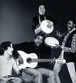 A black and white photo of a man with an acoustic guitar, surrounded by three other men holding percussion instruments