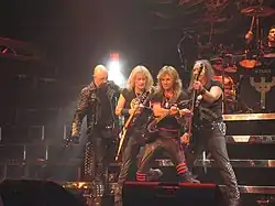 Four men standing next to one another on a stage, three of which are holding guitars. All four men are wearing black clothing, and some of the articles of clothing are studded.