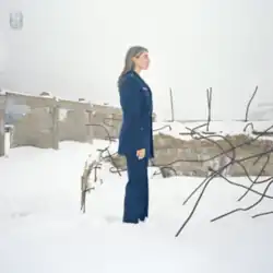Malone wearing a blue suit standing in ankle-deep snow surrounded by dilapidated concrete structures
