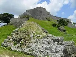A photo of a large grey castle on a grassy hill dotted by stone ruins and small trees.