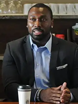 Kerry Carter sits at a table. He's dressed in a dark sport coat and is wearing a blue oxford shirt, unbuttoned at the collar.