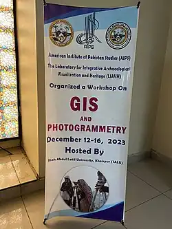 banner for a December 12–16, 2023 Photogrammetry and GIS Workshop. At the top there are three seals: the first being the logo for Shah Abdul Latif University, the second being the logo for the American Institute of Pakistan Studies, and the third a seal for the Office of Research Innovation and Commercialization. At the bottom there is a photo of archaeologist Uzma Rizvi in the field looking through a lens.