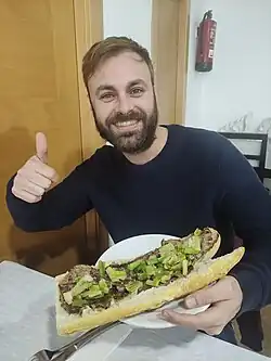 A smiling man holding a long sandwich showing meat and green peppers