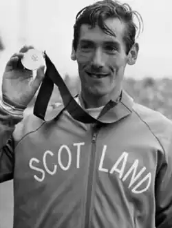 A slim athlete with short hair and moustache wearing a tracksuit top marked "SCOTLAND" smiling and holding up a gold medal
