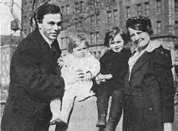 A man and woman standing, each holding a small child, outdoors in winter clothing