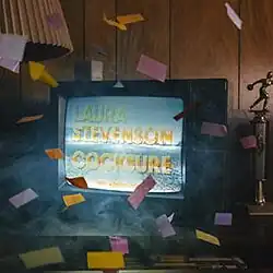A television set in a living room, glowing with blue-gray static and the artist's and album's names in yellow block letters. Multicolored confetti is falling around the room.