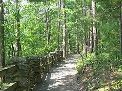 A gravel path through a mixed forest of deciduous and conifer trees, with a rail fence supported by stone pillars left of the path